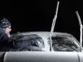 Man skrapar isfri bilruta i mörker. Vindrutetorkarna är täckta av frost.