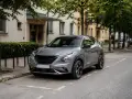 Grå Nissan Juke parkerad längs en gata, med en byggnad och träd i bakgrunden. Kanske ett problem med bilen?