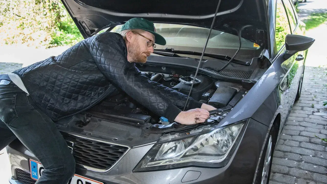 Man med keps och glasögon mekar med bilens motor, kanske byter en h7 led-lampa.