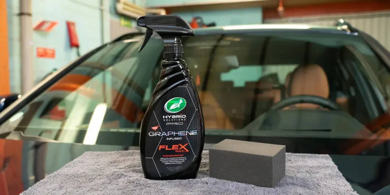 En flaska med "Graphene Flex Wax" och en svamp ligger på en grå trasa framför en bil.