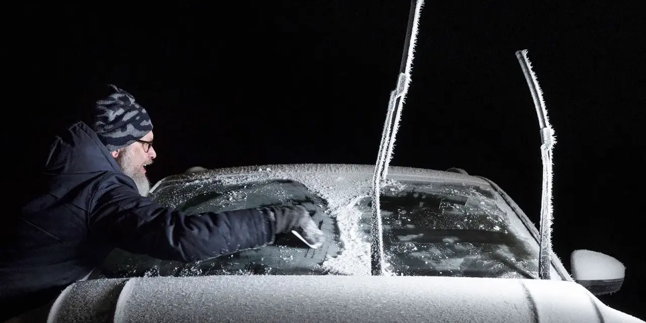 Man skrapar isfri bilruta i mörker. Vindrutetorkarna är täckta av frost.