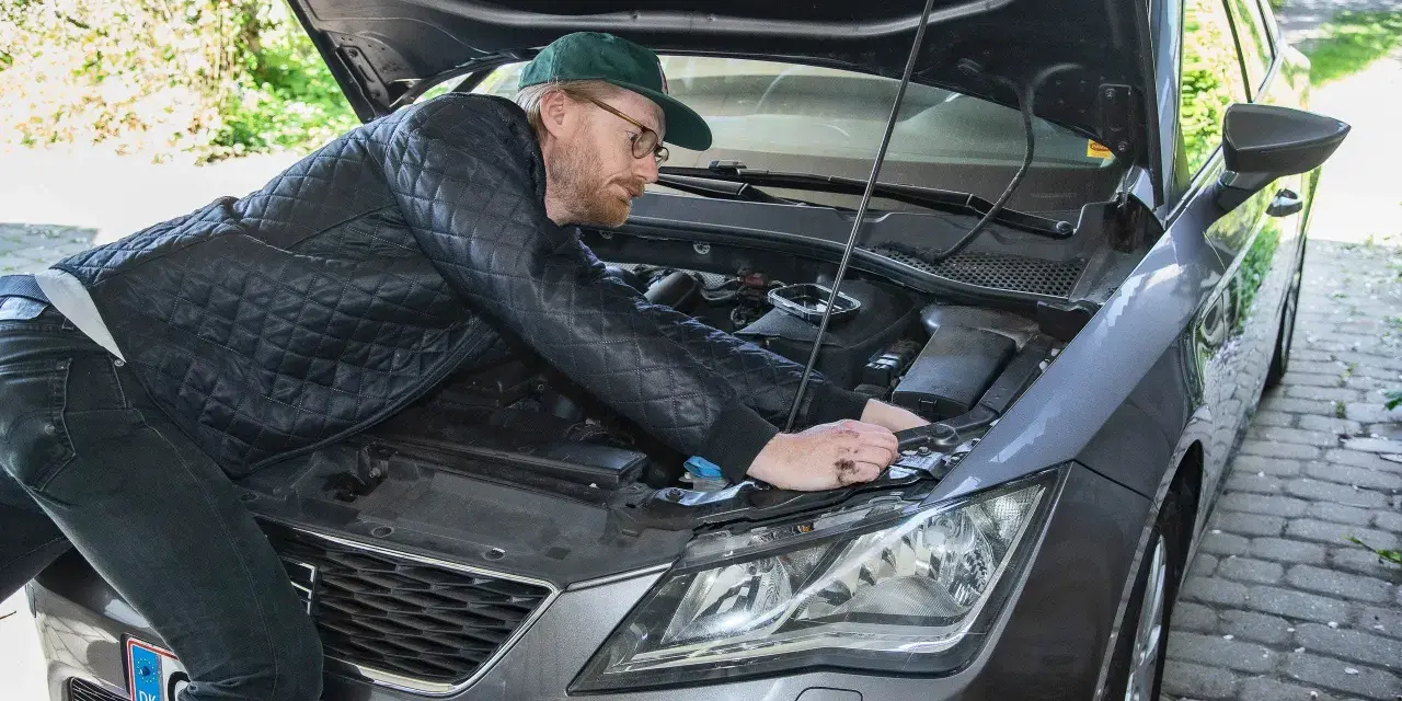 Man med keps och glasögon mekar med bilens motor, kanske byter en h7 led-lampa.