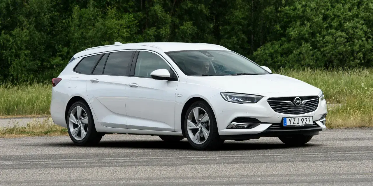 Vit Opel Insignia kombi parkerad vid en grönskande skog. En förare syns i bilen, kanske funderar på ett opel insignia problem.