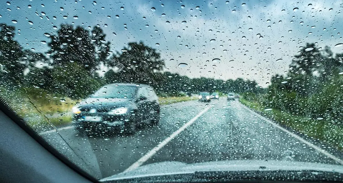 Regndroppar på vindrutan. Bilar kör på en väg genom ett trädbevuxet landskap. Glaco-behandlingen håller sikten klar.