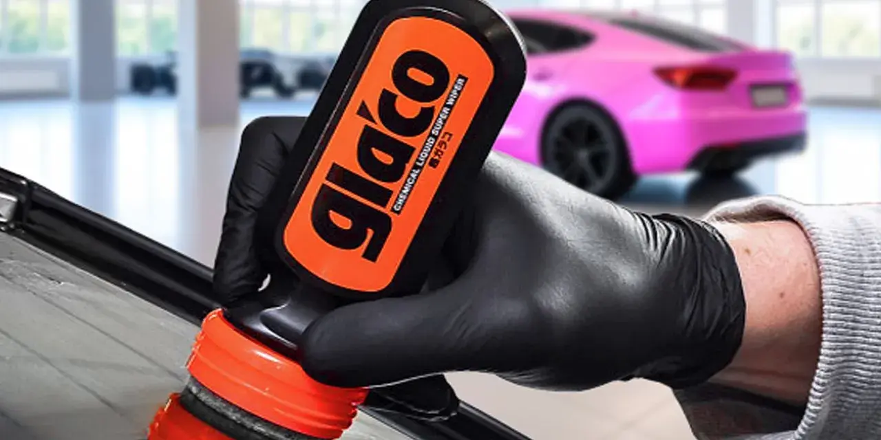 En hand med svart handske applicerar glasförsegling från en Glaco-flaska på en bilruta. En rosa bil syns i bakgrunden.