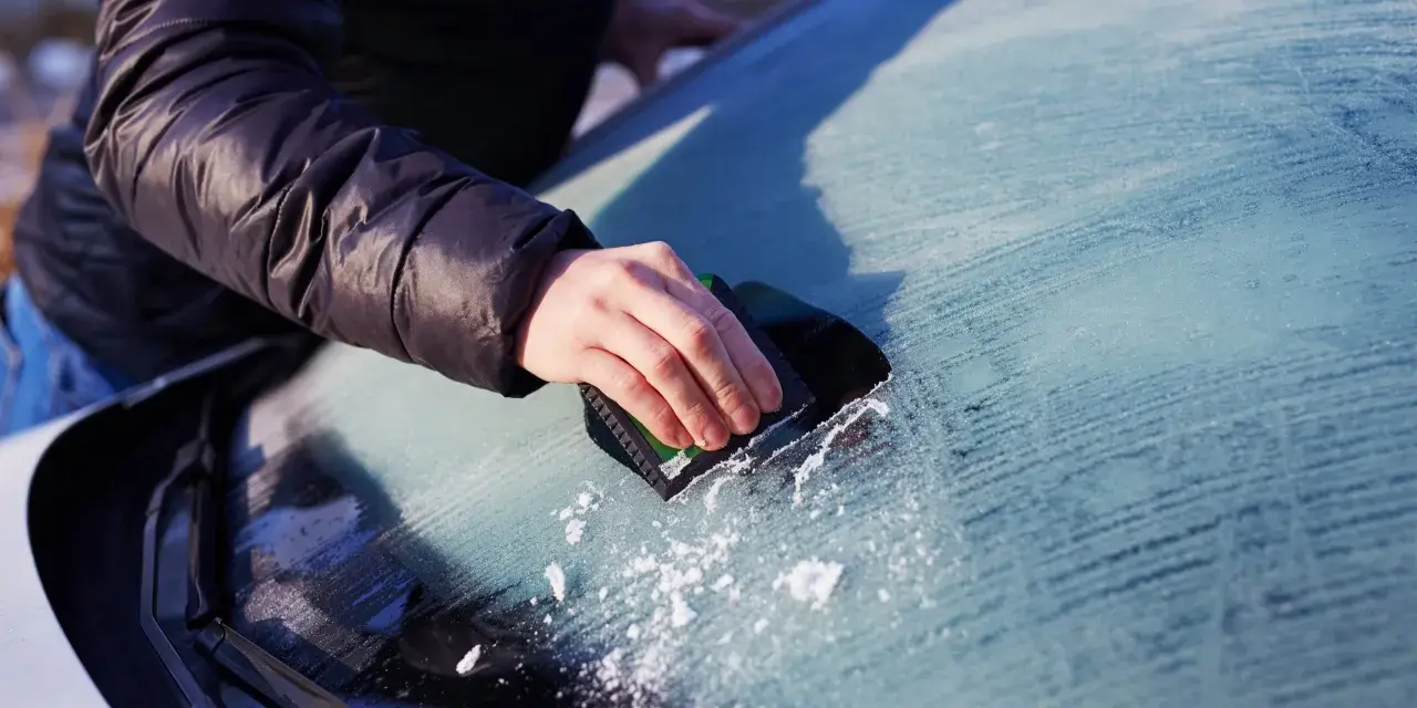 En hand skrapar isfria bilrutor en kall morgon. Snöflingor faller från vindrutan.