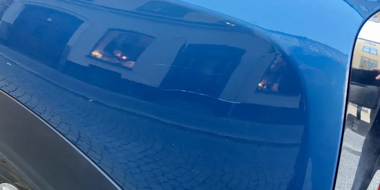 Blå bilskärm med reflektioner av byggnader och kullersten. En repa syns, kanske från kalk borttagning.