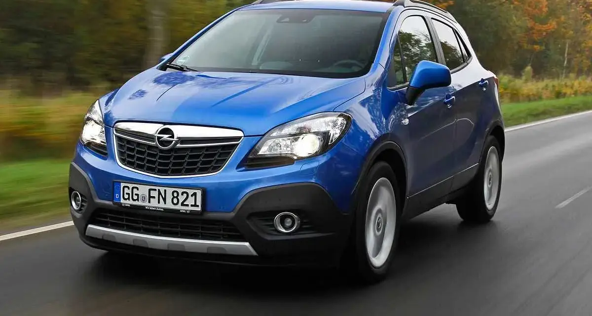 Blå Opel Mokka kör på en väg, kanske med ett opel mokka problem. Träd i höstfärger syns i bakgrunden.