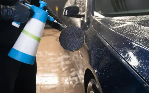 En person med svarta handskar sprutar skum på en mörk metallicbil med en blå och vit sprayflaska. Bilvax metallic ger en glansig finish.