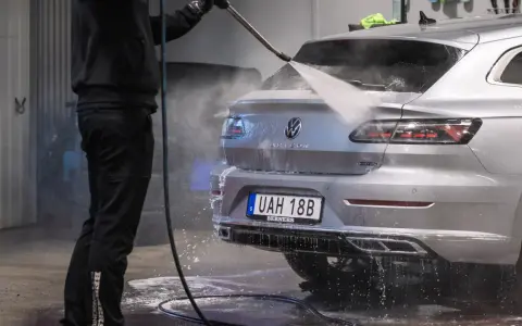 En person tvättar en silverfärgad bil med högtryckstvätt. Frågan "får man tvätta bilen hemma" dyker upp när man ser detta.