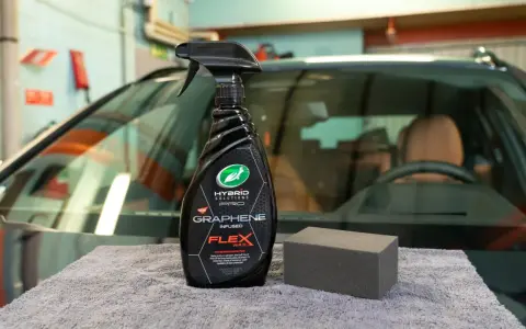 En flaska med "Graphene Flex Wax" och en svamp ligger på en grå trasa framför en bil.