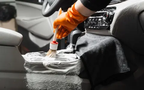 En hand med orange handske använder en **detail brush** för att rengöra bilens mittkonsol med skum.