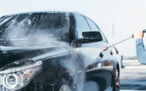 Man med t-grön tröja tvättar en svart bil med högtryckstvätt. Vatten och skum sprutar över bilen.