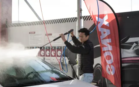 Man using a pressure washer on a car windshield, with a SONAX banner in the background.