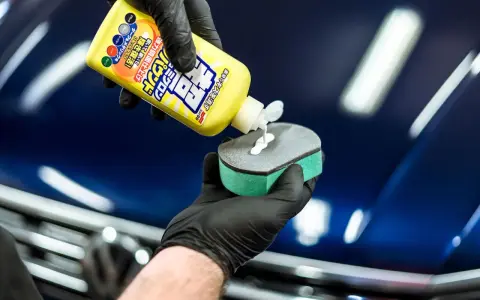 En hand med svart handske applicerar en vit kräm från en gul flaska, märkt Soft 99, på en svamp för bilvård.