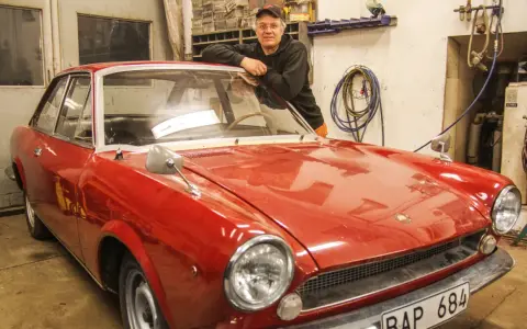Man lutar sig mot en röd klassisk bil i garage. Lite rost syns på bilens skärmkanter.