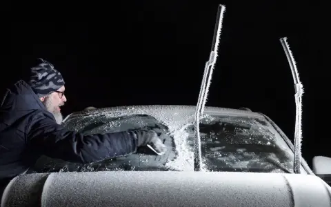 Man skrapar isfri bilruta i mörker. Vindrutetorkarna är täckta av frost.
