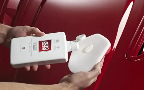 Händer applicerar Autoglym Super Resin Polish på en blank röd bil med en svamp.