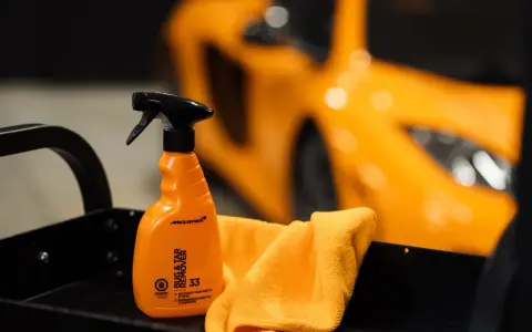 En orange sprayflaska för att ta bort limrester och insekter, bredvid en orange mikrofiberduk. En sportbil i bakgrunden.