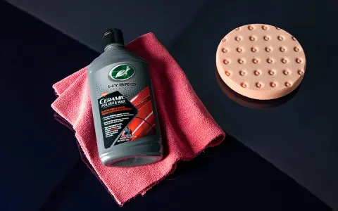 En flaska Turtle Wax Hybrid Solutions Ceramic Polish & Wax, en rosa mikrofiberduk och en polertrissa ligger på en mörkblå yta.