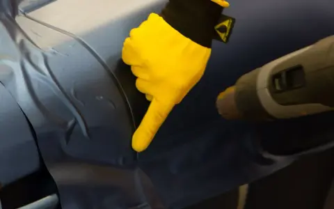 Gul handske pekar på bilens yta där foliering ska tas bort med värmepistol.
