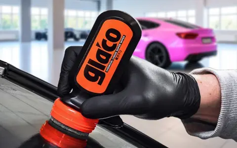 En hand med svart handske applicerar glasförsegling från en Glaco-flaska på en bilruta. En rosa bil syns i bakgrunden.