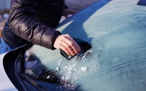 En hand skrapar isfria bilrutor en kall morgon. Snöflingor faller från vindrutan.