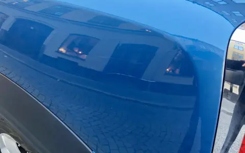 Blå bilskärm med reflektioner av byggnader och kullersten. En repa syns, kanske från kalk borttagning.
