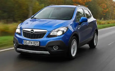 Blå Opel Mokka kör på en väg, kanske med ett opel mokka problem. Träd i höstfärger syns i bakgrunden.