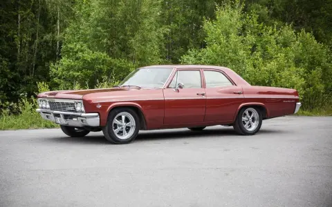 Röd Chevrolet Impala från 1965 med en tung cut, parkerad vid en skogsdunge.