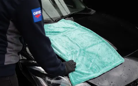 Person i handskar lägger en turkos mikrofiberduk på en svart bilhuv. Duken är från the rag company.