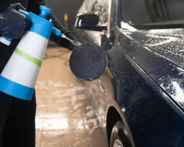 En person med svarta handskar sprutar skum på en mörk metallicbil med en blå och vit sprayflaska. Bilvax metallic ger en glansig finish.