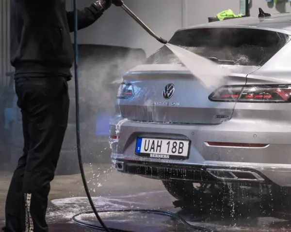 En person tvättar en silverfärgad bil med högtryckstvätt. Frågan "får man tvätta bilen hemma" dyker upp när man ser detta.