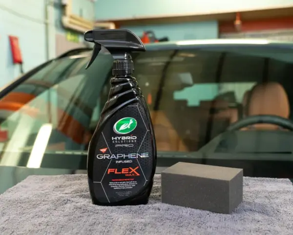 En flaska med "Graphene Flex Wax" och en svamp ligger på en grå trasa framför en bil.