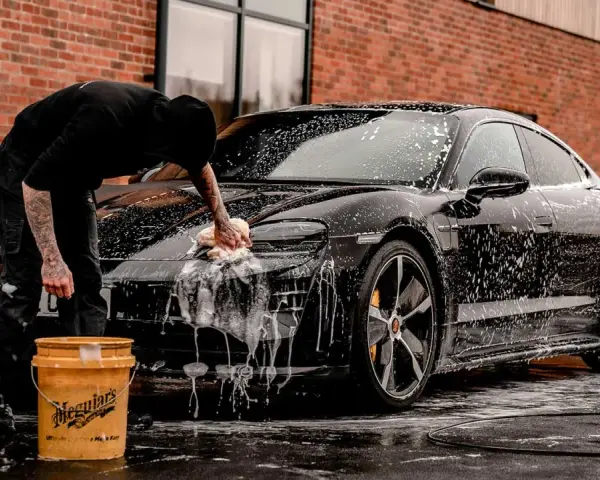 Person tvättar en svart bil med Autoglym-produkter. Två gula hinkar står bredvid.