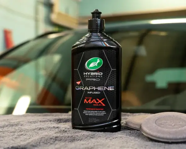 En flaska Turtle Wax Hybrid Solutions Pro Graphene Infused To The Max Wax står på en grå trasa bredvid två appliceringsdynor.