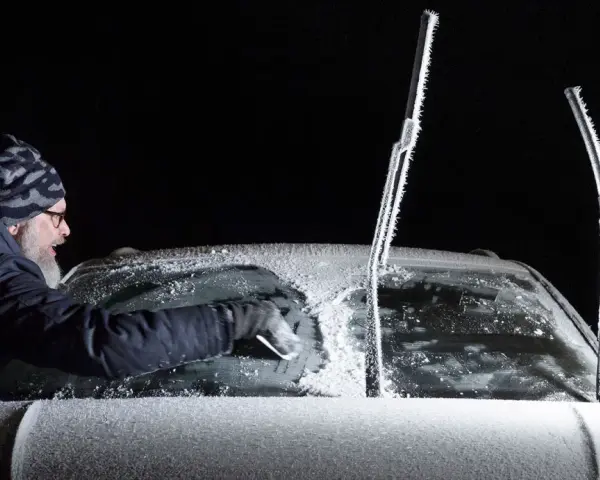 Man skrapar isfri bilruta i mörker. Vindrutetorkarna är täckta av frost.