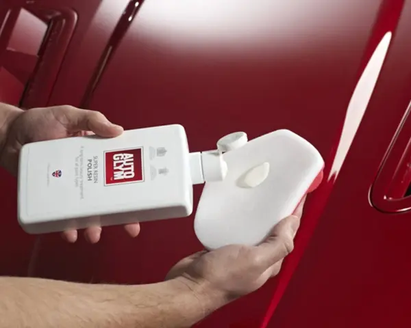 Händer applicerar Autoglym Super Resin Polish på en blank röd bil med en svamp.