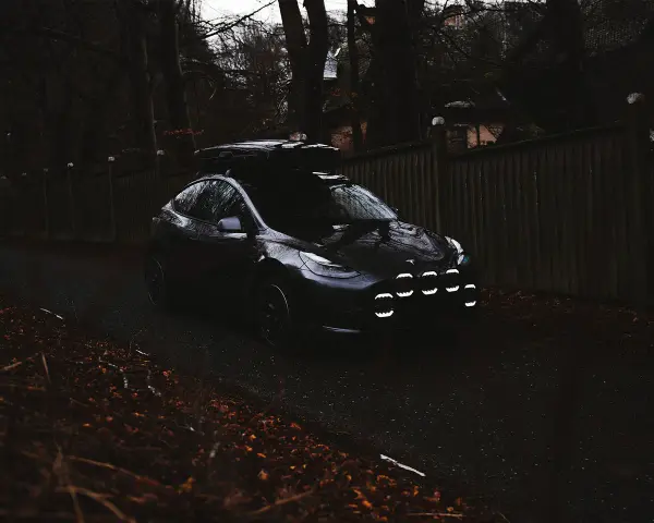 Mörk Tesla med takbox och extraljusstag redo för äventyr längs en höstfärgad väg.