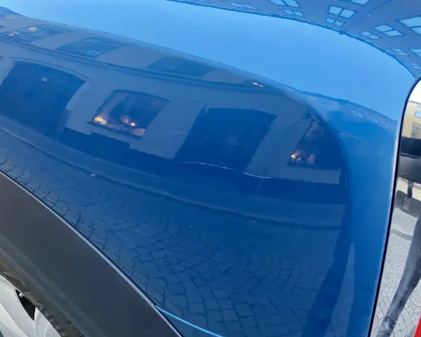 Blå bilskärm med reflektioner av byggnader och kullersten. En repa syns, kanske från kalk borttagning.