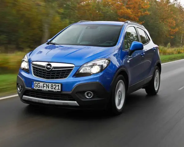 Blå Opel Mokka kör på en väg, kanske med ett opel mokka problem. Träd i höstfärger syns i bakgrunden.