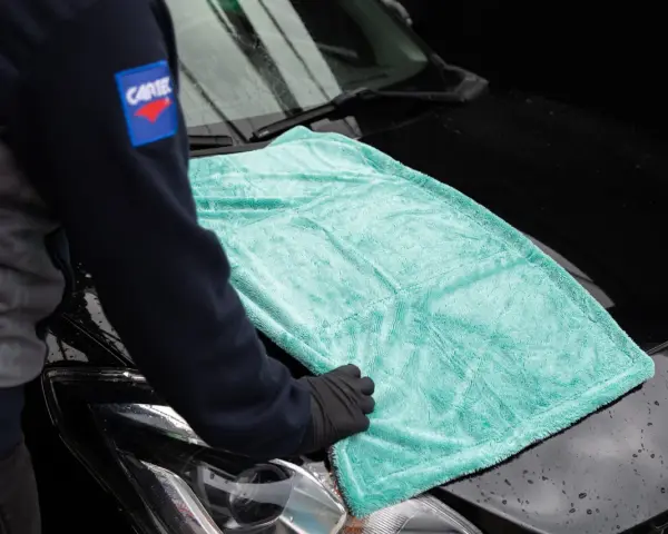 Person i handskar lägger en turkos mikrofiberduk på en svart bilhuv. Duken är från the rag company.