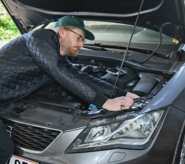 Man med keps och glasögon mekar med bilens motor, kanske byter en h7 led-lampa.