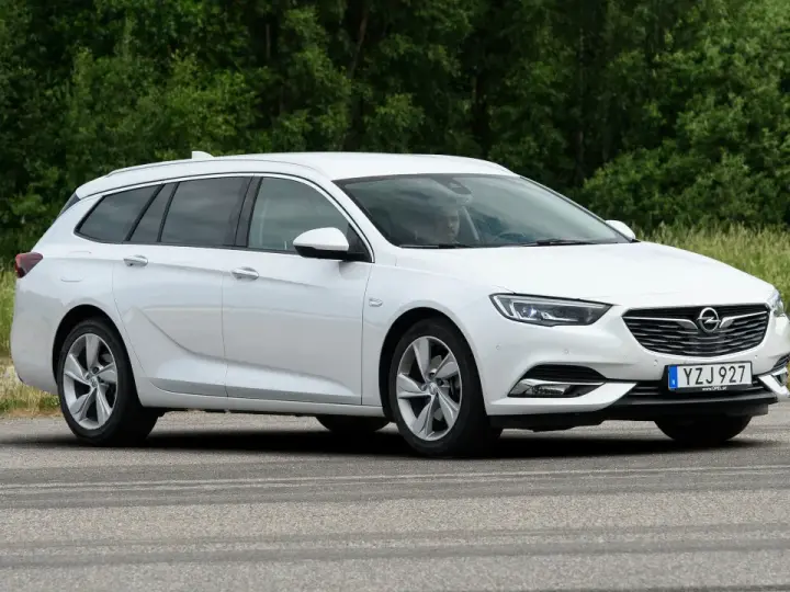 Vit Opel Insignia kombi parkerad vid en grönskande skog. En förare syns i bilen, kanske funderar på ett opel insignia problem.