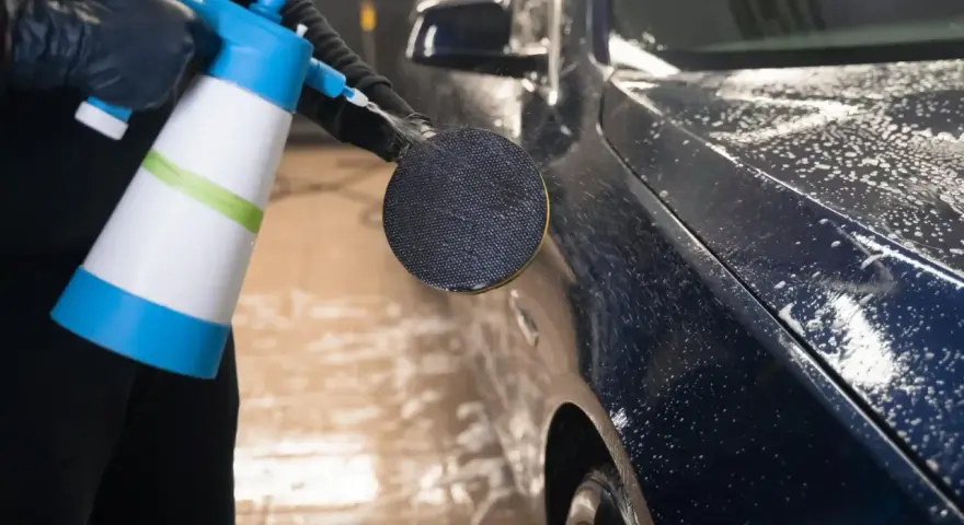 En person med svarta handskar sprutar skum på en mörk metallicbil med en blå och vit sprayflaska. Bilvax metallic ger en glansig finish.