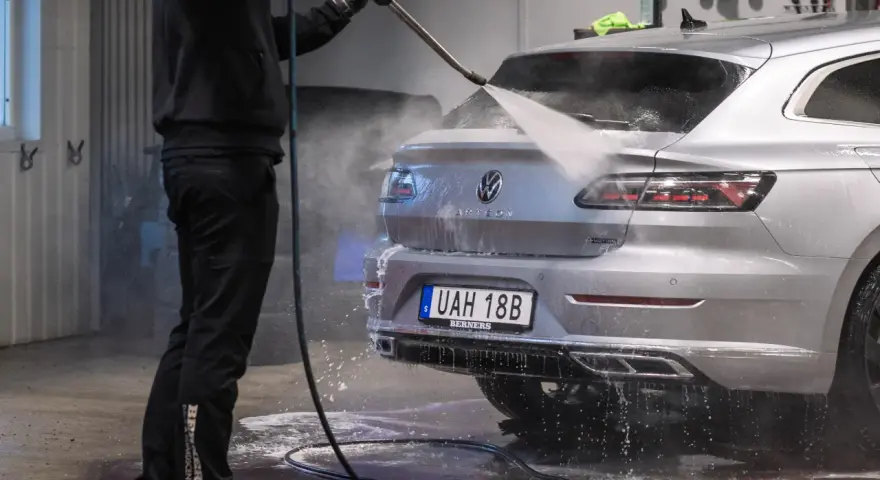 En person tvättar en silverfärgad bil med högtryckstvätt. Frågan "får man tvätta bilen hemma" dyker upp när man ser detta.