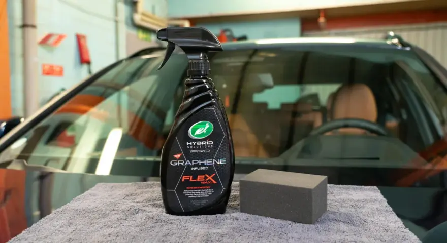 En flaska med "Graphene Flex Wax" och en svamp ligger på en grå trasa framför en bil.