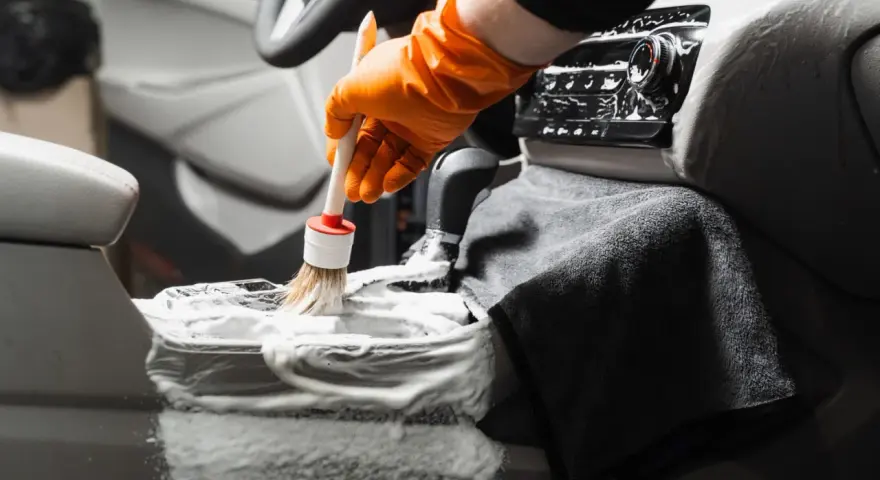 En hand med orange handske använder en **detail brush** för att rengöra bilens mittkonsol med skum.