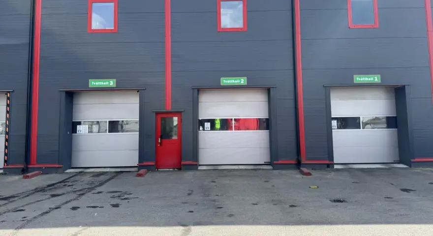 Grå fasad med röda detaljer och tre portar märkta Tvätthall 1, 2 och 3. En röd dörr finns mellan portarna.