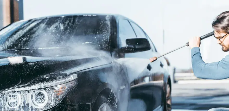 Man med t-grön tröja tvättar en svart bil med högtryckstvätt. Vatten och skum sprutar över bilen.